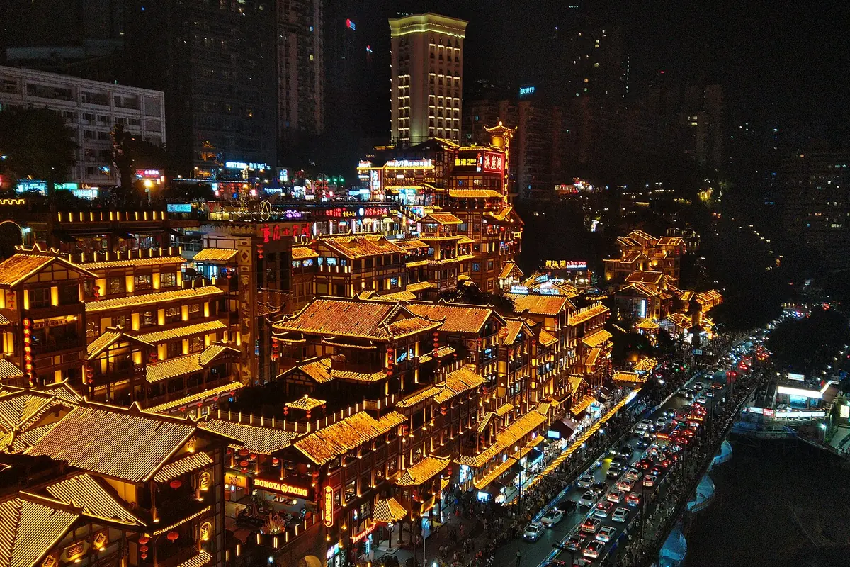 Tiers of gold-lit stilt-style buildings climbing a hillside above a road at night in Chongqing